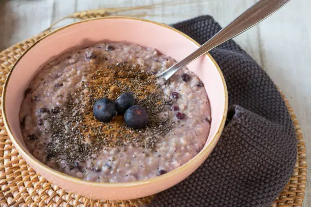 fresh cooked and homemade porridge with blueberries, chia seeds and cinnamon served in a pale pink bowl on a wooden kitchen table from above
