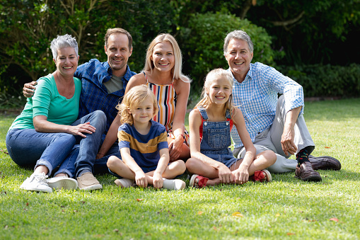 Portrait of caucasian parents, grandparents and grandchildren sitting in garden smiling to camera. happy three generation family spending time together at home.