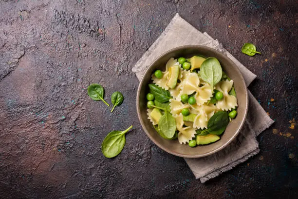 Top view of tasty vegetarian farfalle pasta with green peas, avocado and spinach leaves on brown concrete background