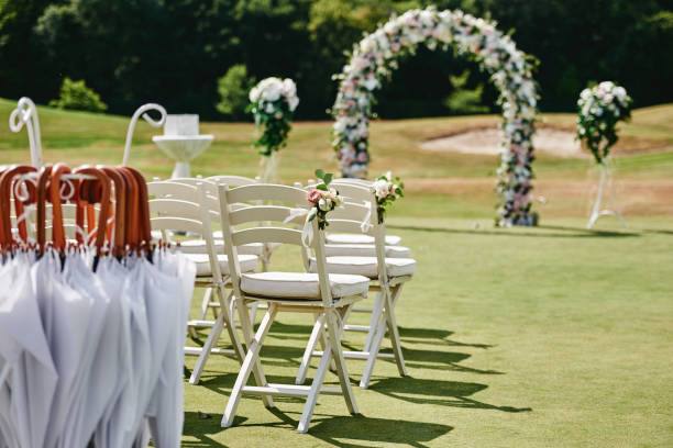 屋外のアーチウェイの両側にバラの花を持つ白い木製の椅子、コピースペース。ゴルフコースでの結婚式のために準備されたゲストのための空の椅子 - スポーツ施設 ストックフォトと画像