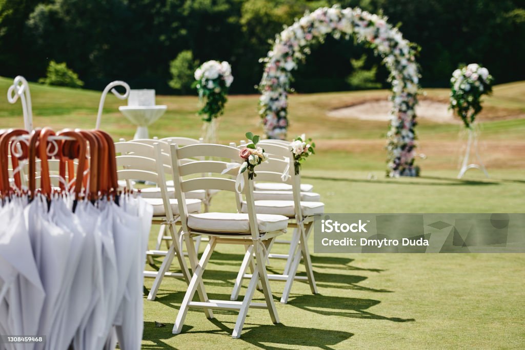 White wooden chairs with rose flowers on each side of archway outdoors, copy space. Empty chairs for guests prepared for wedding ceremony on golf course Wedding Stock Photo