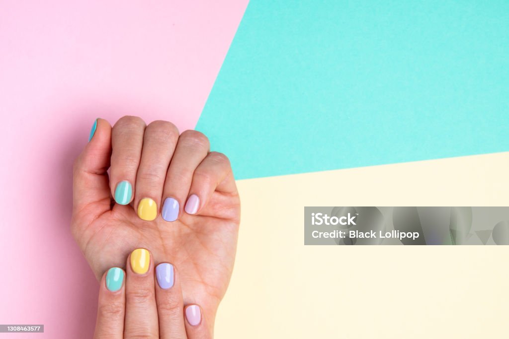 Hands with pastel nail polish on multicolored background. Bright trendy female manicure in funky spring colors. Woman's hands with pastel manicure on yellow, pink and blue background. Beautiful multi colour spring Easter manicure. A lot of space for text. Fingernail Stock Photo