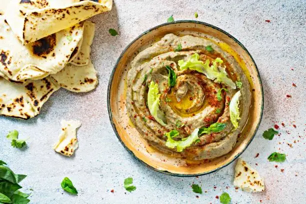 Photo of An oriental dish of baked eggplant babaganush (eggplant puree) with spices, herbs, lettuce and oriental flatbreads on a light background.