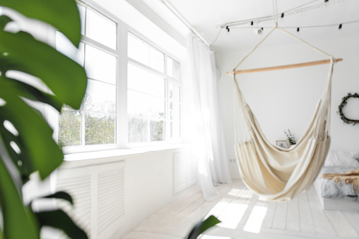 Stylish, trendy interior in Scandinavian style. Large wooden window sees white macrame hammock against the backdrop of green monstera, bed on the floor. Garland of light bulbs hangs from the ceiling.