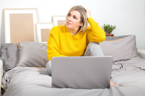 A pensive focused woman is sitting on the bed, working with a laptop and looking out the window, pondering a strategy. Solving the problem. Work from home, freelance. Color trend 2021 A pensive focused woman is sitting on the bed, working with a laptop and looking out the window, pondering a strategy. Solving the problem. Work from home, freelance. Color trend 2021. food sample cups stock pictures, royalty-free photos & images