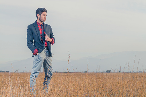 Young latin chilean handsome Attractive Elegant business fashion young man with nice smile in casual clothing modeling and posing outdoors, wearing a black jacket, grey pants, red square shirt and black tie; Standing around to autumnal wheat field, on back the  Chilean Patagonia Mountains in an autumn cloudy winter cold afternoon.\n\nFull body length shoot. Plenty Space for copy text.