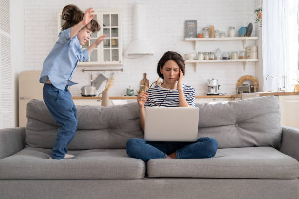 zmęczona mama siedzi na kanapie, pracuje na laptopie w domu, nadpobudliwe małe dziecko skacze przyciągając uwagę. - working mother child emotional stress mother zdjęcia i obrazy z banku zdjęć