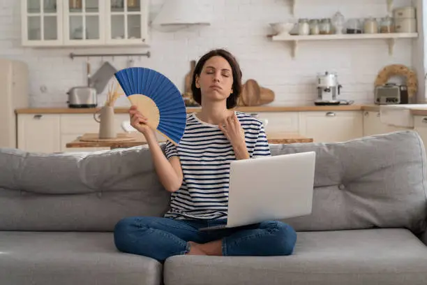 Exhausted young woman suffering from heatstroke flat without air-conditioner, waving blue fan, sitting on couch at home, working on laptop computer. Overheating, high temperature, hot summer weather.