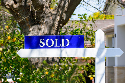 Sold real estate sign on wooden post in the front of a house. Blurred green landscaping background.