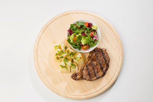 Barbecue meal on wooden dish: Grilled meat, cassava with vegetable dressing and mixed salad with tomato and variation of lettuce.