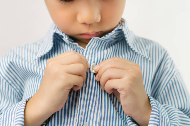 Cute Asian boy getting dressed for school, buttoning long sleeved shirt with concentration and happiness to go back to school Close up of studio shot of small cute elementary age Asian boy learning and practicing buttoning shirt. Child sensory motor, muscle and sensory perception development in Montessori education. Self confidence and independence growth. Independence in Daily Activities:  in kids stock pictures, royalty-free photos & images