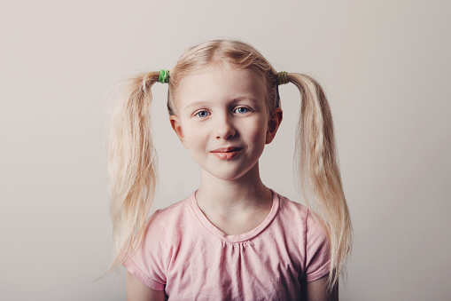Closeup portrait of cute smiling blonde Caucasian preschool girl in pink t-shirt on light background. Child with long pig tails hair posing looking in camera. Kid expressing positive emotions.
