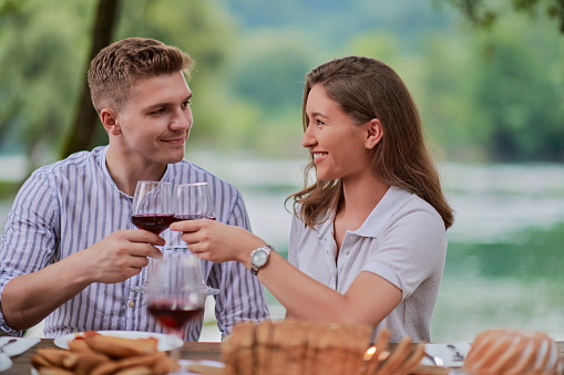 young happy couple toasting red wine glass while having picnic french dinner party outdoor during summer holiday vacation near the river at beautiful nature