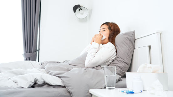 Young Asian Woman with seasonal infections Cold Blowing Her Nose and sneezing into Tissue with headache lying under the blanket in bed with high fever and a flu.