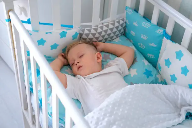 A charming boy sleeps in a baby white crib. A small child sleeps in a crib on blue bedding. Sleep workout concept.