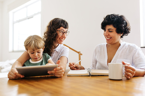 Lesbian couple looking at each other and smiling with son using tablet pc at home. Female couple smiling while their son using digital tablet.