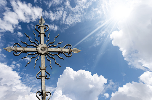 Closeup of a wrought iron religious cross on beautiful blue sky with clouds and sun rays. Veneto, Italy, Europe.