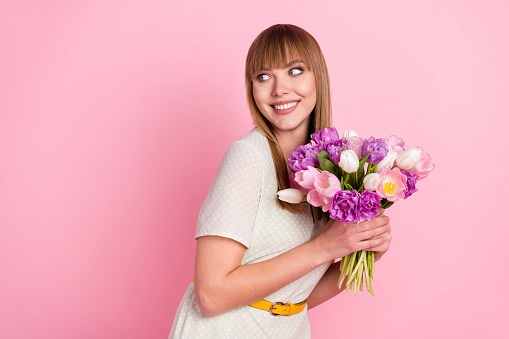 Portrait of attractive cheerful glad girl holding in hands flowers looking aside copy space isolated over pink pastel color background.