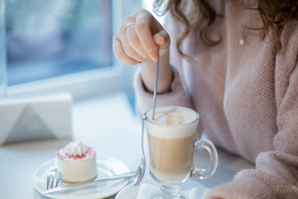 taza de cristal con capuchino y pastel en un plato. una mujer sosteniendo una cuchara larga en tu mano. - women spoon tasting elegance fotografías e imágenes de stock