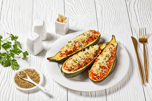 zucchini boats stuffed with ground meat, chopped tomatoes, and cheese on a white platter on a wooden table,  italian cuisine