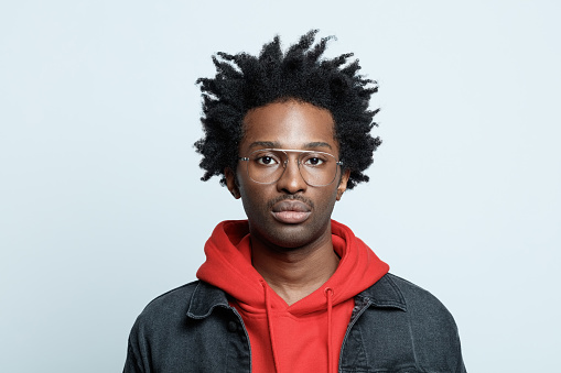 Afro american young man wearing red hoodie, black jeans jacket and glasses, looking at camera. Studio shot on grey background.