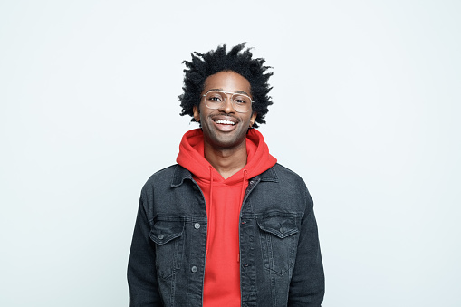 Cheerful afro american young man wearing red hoodie, black jeans jacket and glasses, smiling at camera. Studio shot on grey background.