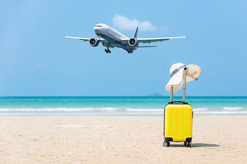 Summer traveling and planning with yellow suitcase luggage with big white hat fashion on the sand beach. Travel in the holiday, airplane and blue sky and beach background. Summer and Travel Concept