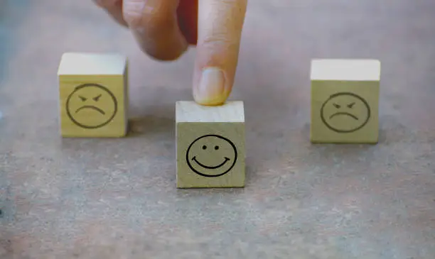 Photo of Customer hand finger choosing happy smiley face on wooden cube. Two blurred sad face icon on wooden cubes in background, Illustration of rating, consumer satisfaction, performance, team building.