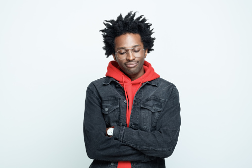 Sad afro american young man wearing red hoodie, black jeans jacket and glasses, looking down with arms crossed. Studio shot on grey background.