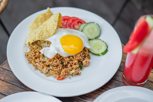 Close-up shot of Indonesian fried rice with sunny side up egg ( nasi goreng ) and slice of cucumber, tomatoes and a cracker. There's a watermelon juice on the side of the plate.