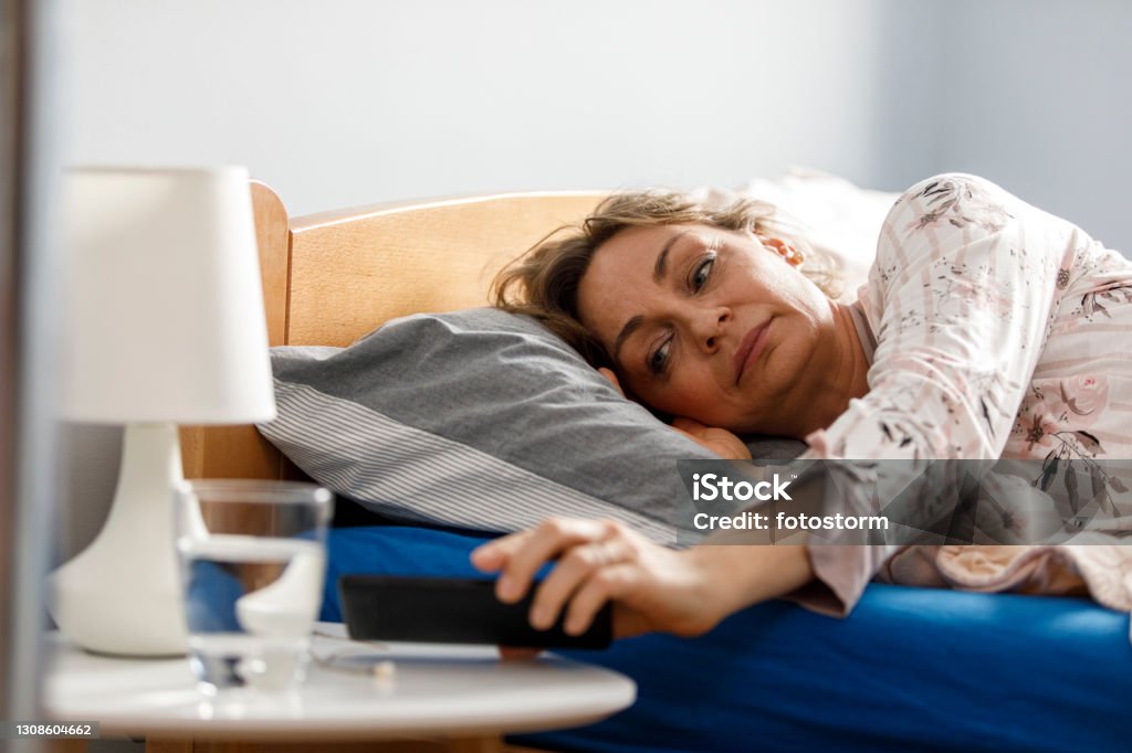 Turning off the alarm clock on her smart phone in the morning Tired mature woman lying in cozy bed in the morning and getting her smart phone to turn off or snooze the alarm clock. Mature Women Stock Photo