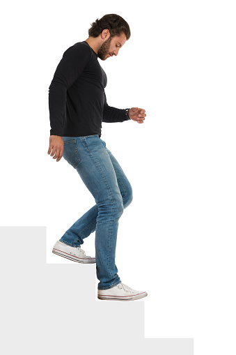 Casual serious young man in jeans, sneakers and black jersey is going down the stairs and looking away. Side view. Full length studio shot isolated on white.