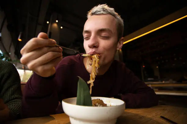 Photo of Satisfied man enjoys food in the restaurant. Blong attractive young man with pierced nose puts pasta into his mouth and closes his eyes with pleasure. Tasty food concept.