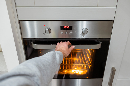 A point of view shot of an unrecognisable mature caucasian woman opening an oven in her kitchen at home. She is pre-heating the oven to bake muffins and bread.