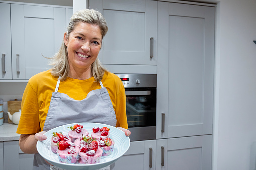 A mature caucasian woman smiling and looking at the camera while she holds a plate of muffins she has just baked and decorated. She is wearing casual clothing and an apron while she stands in her kitchen at home.