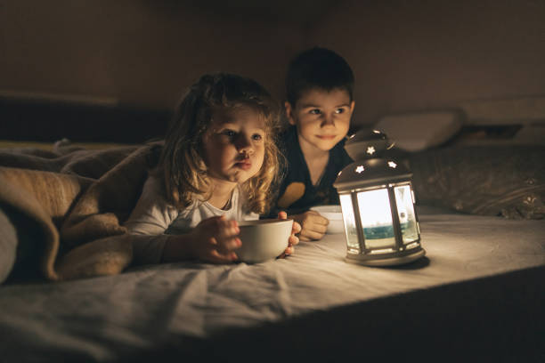 Cute little brother and sister lying on bed under blanket in dark room and eating snacks while glowing rustic lantern lamp