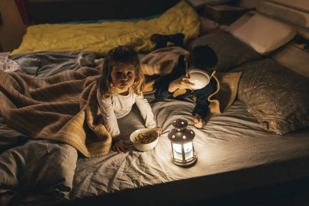 istock Brother and sister lying on bed under blanket in dark room and eating snacks while glowing rustic lantern lamp 1308617833