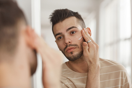 Portrait of young man applying face cream while looking in mirror during morning skincare routine, copy space