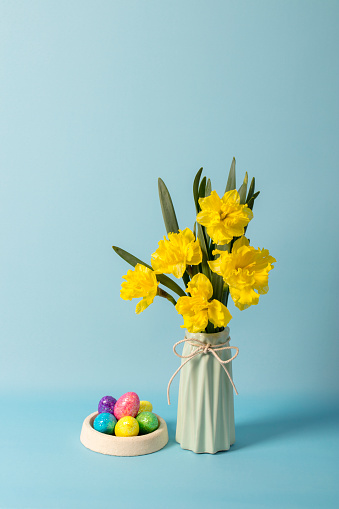 Easter eggs and narcissus (daffodil) flowers in a vase. Blue background. Space for copy.