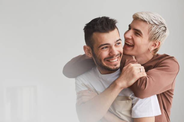 Portrait of Carefree Gay Couple Indoors Candid waist up portrait of carefree gay couple embracing indoors and laughing happily while posing against white, copy space man gay stock pictures, royalty-free photos & images