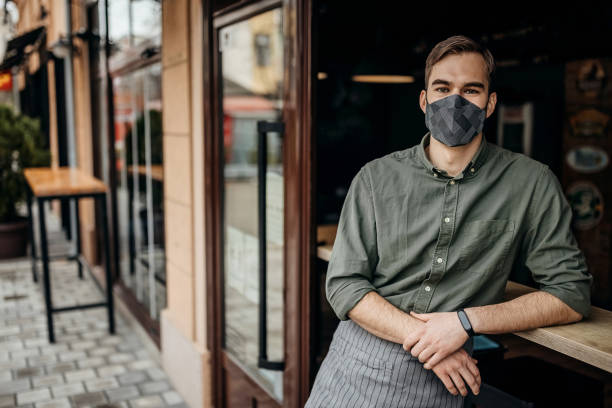 istock Young waiter with face mask posing in front of cafe 1308624733