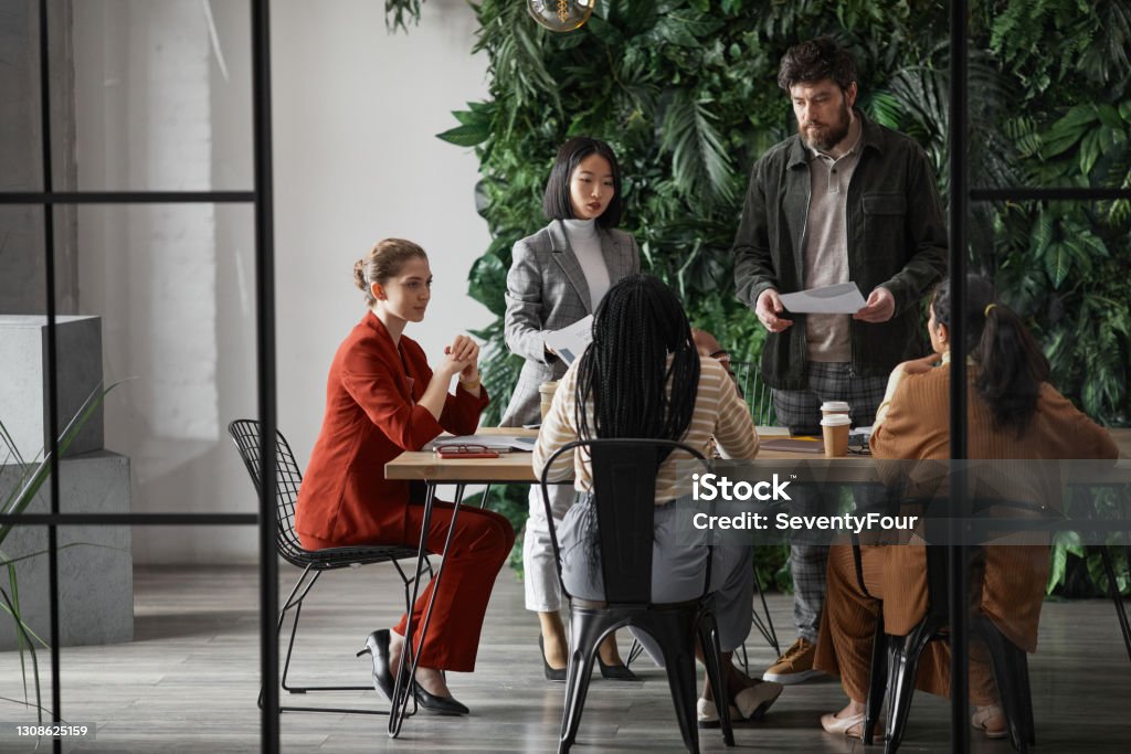 Diverse Business Team in Graphic Office Graphic full length portrait of diverse group of business people meeting at table in modern office interior, copy space Office Stock Photo