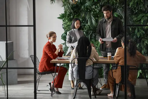 Graphic full length portrait of diverse group of business people meeting at table in modern office interior, copy space