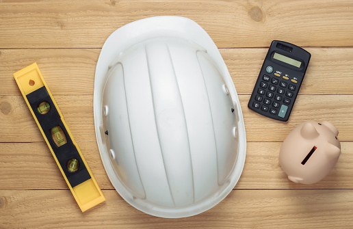 White construction safety helmet with level, piggy bank and calculator on wooden background. The cost of repairing or building house. Top view.