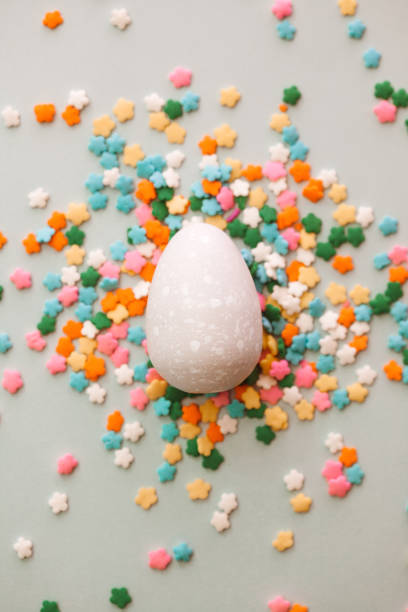 istock White Easter egg on grey background with colourful sprinkles. Yellow, orange, pink, white, blue and green sugar edible decoration  with Easter egg. 1308637941