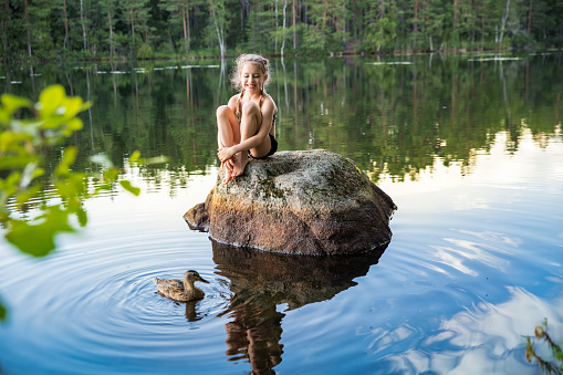 Cute little girl sitting on a rock in lake. Enjoying summer vacation. Child and Nature. Happy isolation concept. Exploring Finland. Scandinavian landscape.