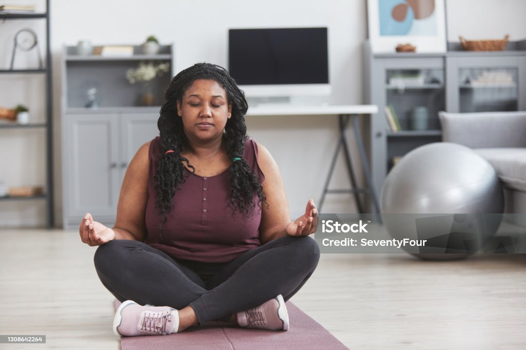 Overweight African American Woman Meditating at Home Front view full length portrait of curvy African American woman meditating at home while sitting on mat and enjoying home yoga practice, copy space Meditating Stock Photo