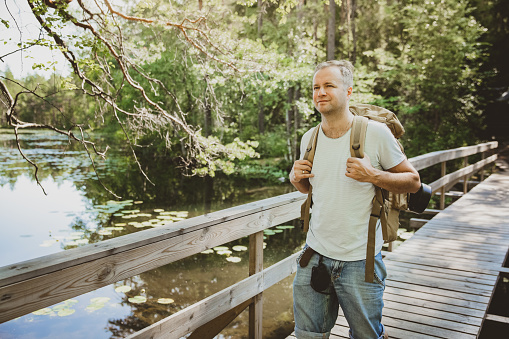Mature man exploring Finnish nature in summer, walking across the bridge. Hiker with big backpack traveling in forests. Summer Scandinavian landscape of lakes and woods.
