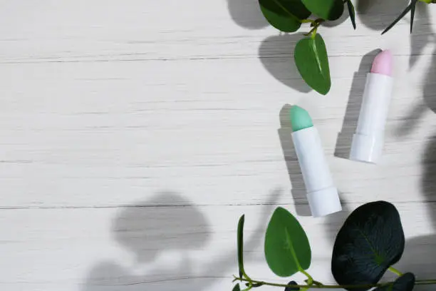 Photo of White tubes of pink and green lip balm with aloe, against dryness, to protect and nourish lips, on a white wooden background with leaves and harsh shadows. Cosmetics for the spring-summer season.