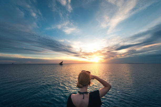 Rear view on young woman watching traditional fishing boat (dhow) at sunset (Zanzibar, Tanzania, Africa).